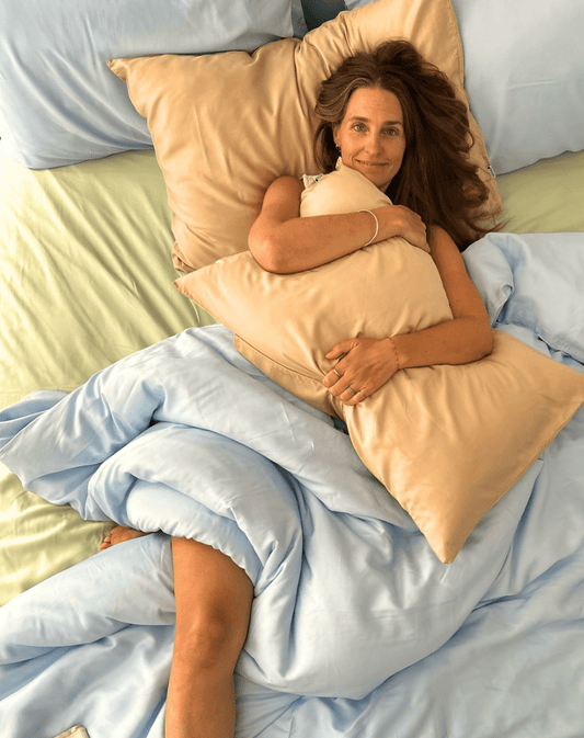 Femme au lit tenant une parure de draps en coton pêche avec couette bleue, drap vert clair visible dessous