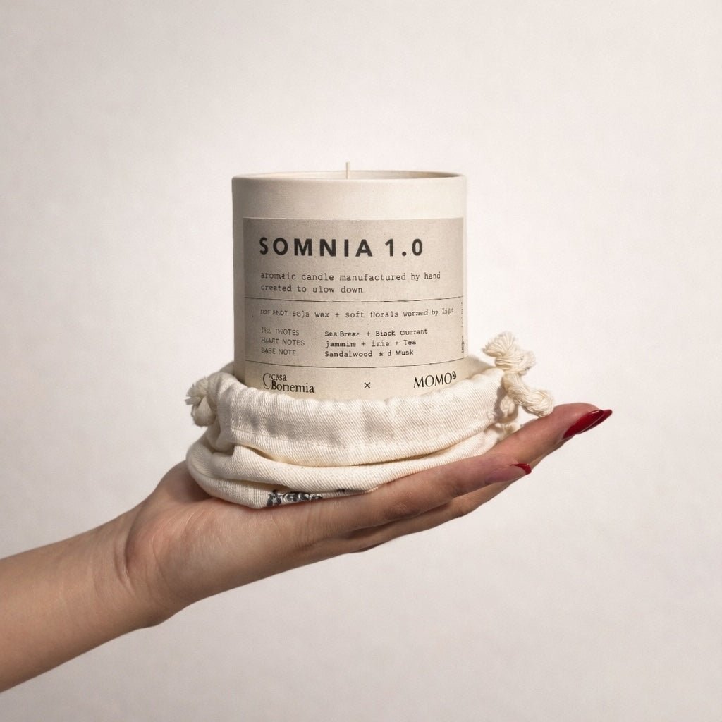 Main tenant une bougie aromatique Somnia 1.0 couleur crème enveloppée dans du coton naturel sur fond blanc