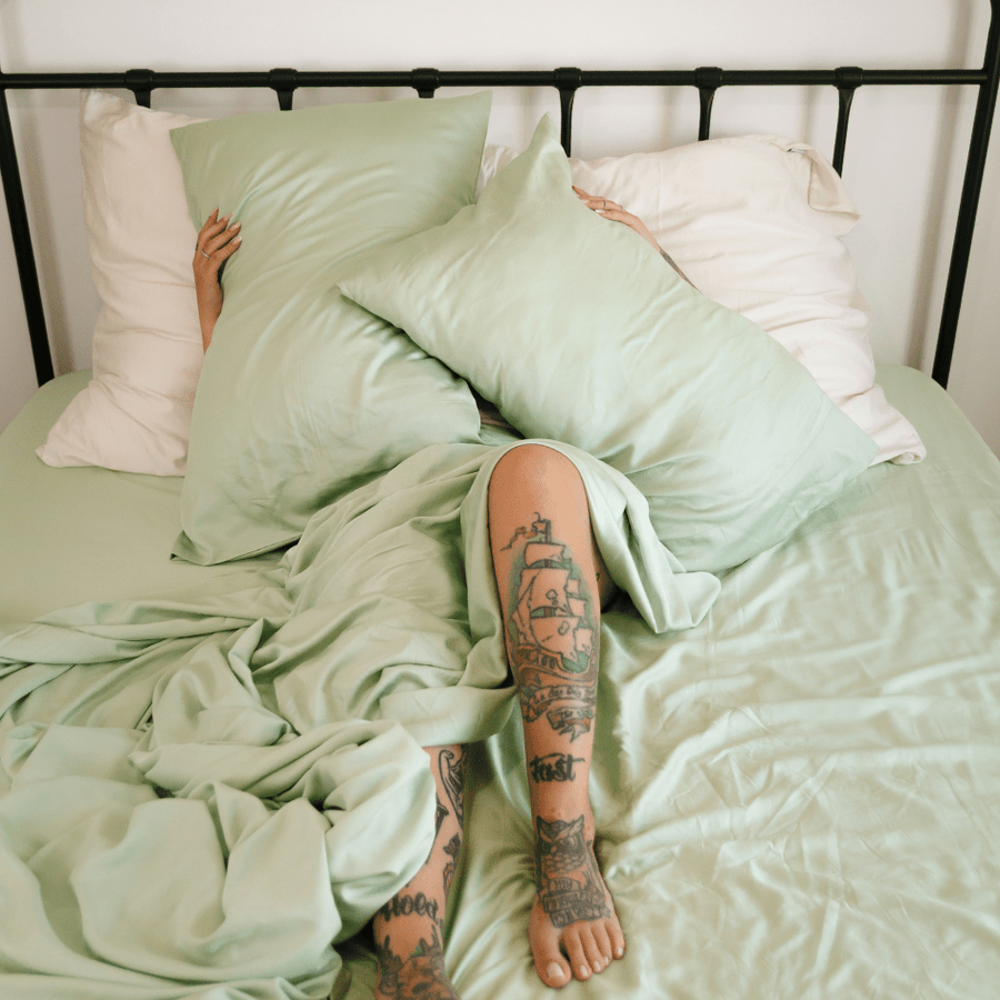 Personne allongée au lit portant une parure de lit en bambou vert et blanc avec des tatouages visibles sur les jambes