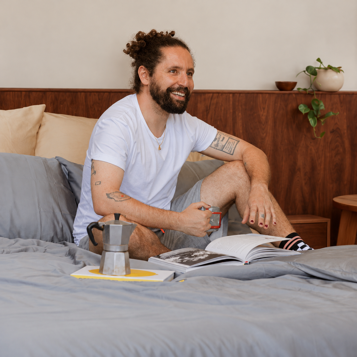 Man in bed with gray cotton bedding reading magazine with moka coffee pot