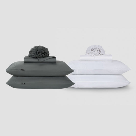 Two Momo Cotton sheet sets displayed side-by-side, one charcoal gray and one white, each stacked with pillows and rolled